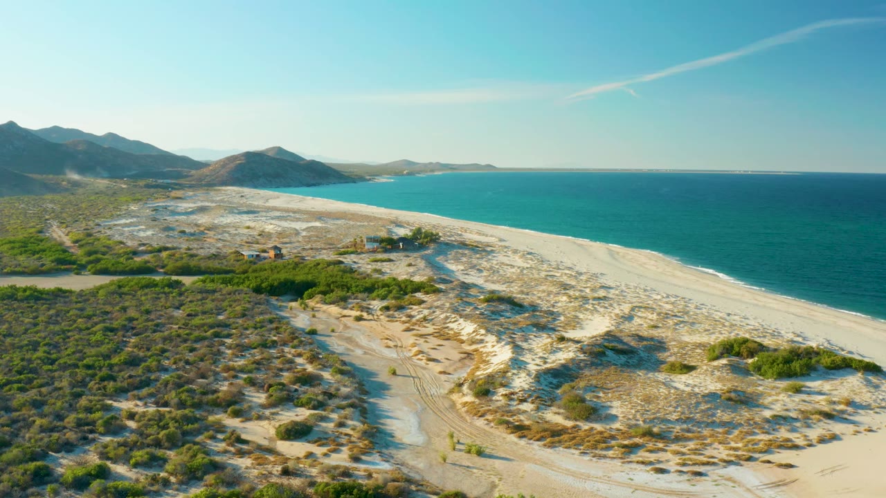 Aerial view in Las Barracas with the shoreline stretching diagonally beside turquoise water, scrubland filling the left foreground, distant mountains behind the dunes, and no visible people.