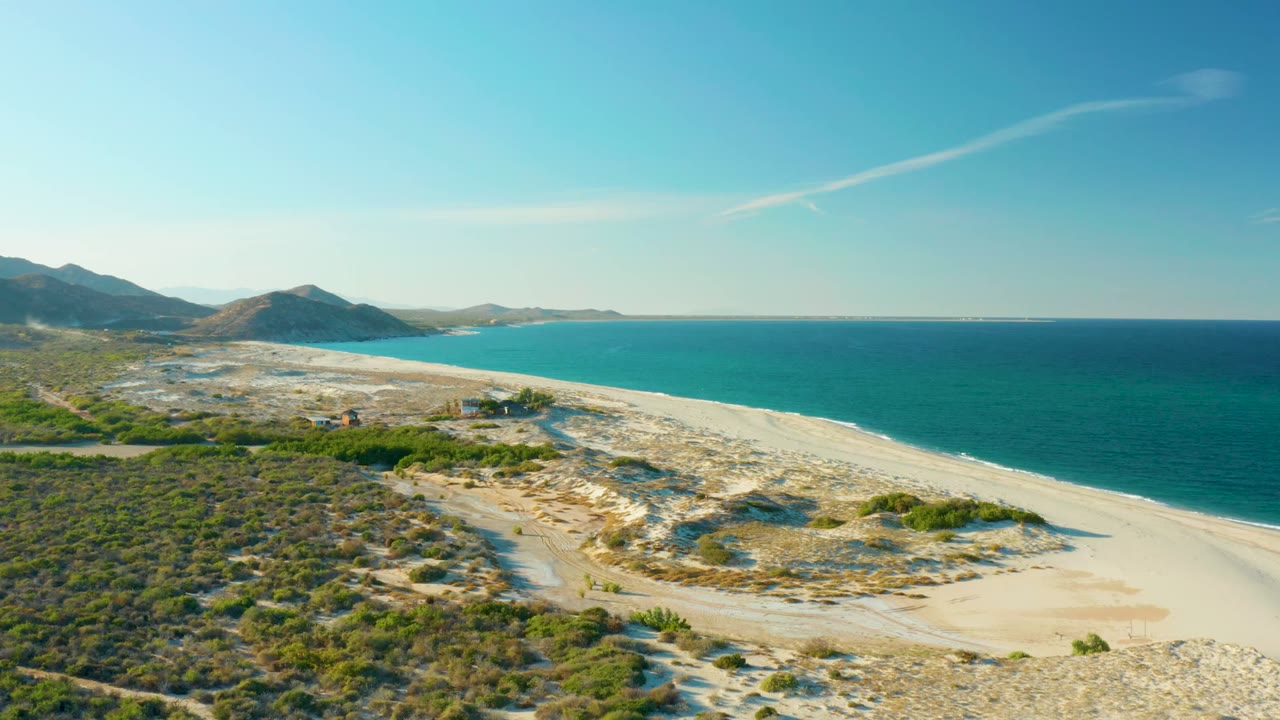 Aerial frame over Las Barracas coastline showing scrubland and pale sand in the foreground, blue ocean at right, distant mountains at left, and no visible people.