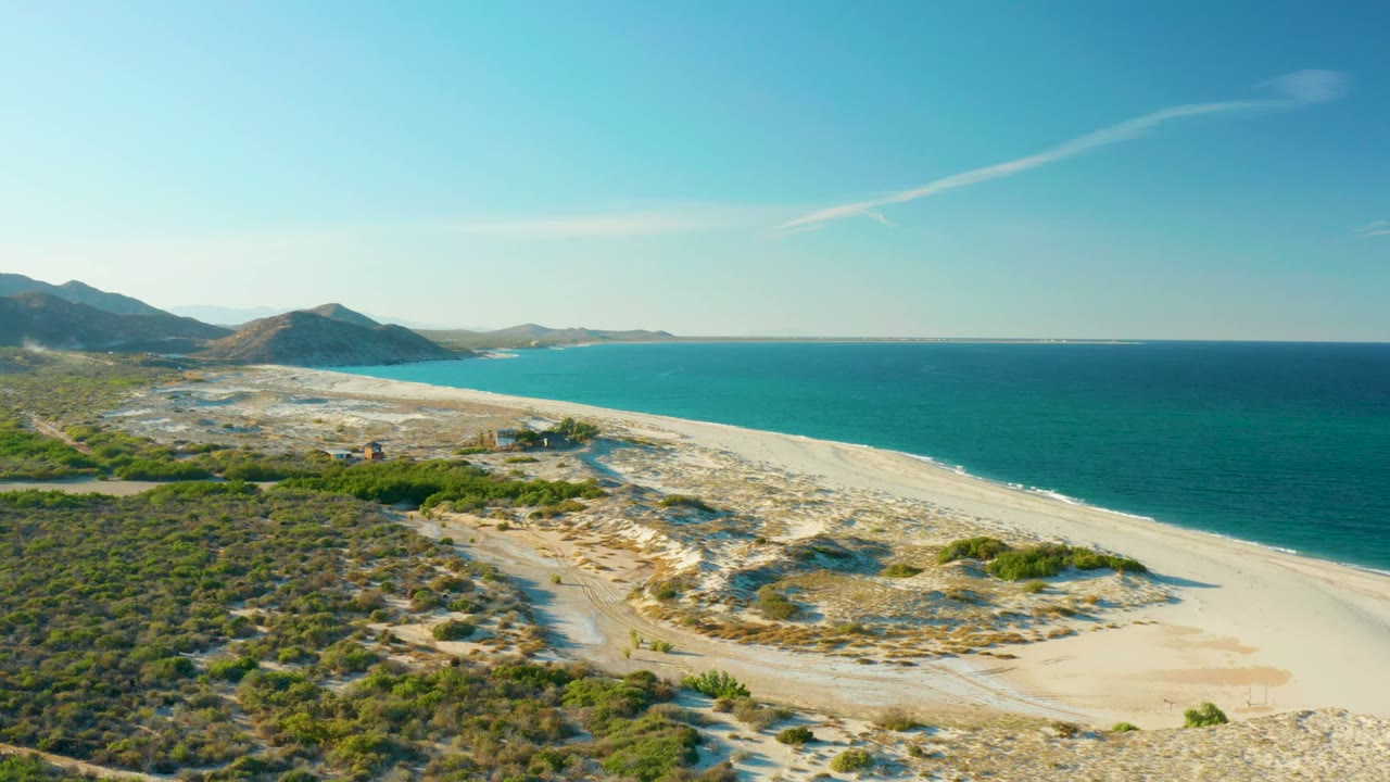 Wide drone view of beach dunes, turquoise water, scrub vegetation, and low mountains.