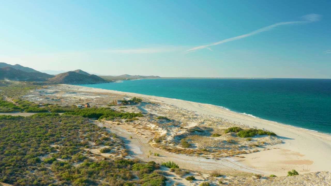 Aerial view of the Las Barracas beach with bright sandbars and dune texture in the center, deep blue water along the right edge, mountains beyond, and no visible people.