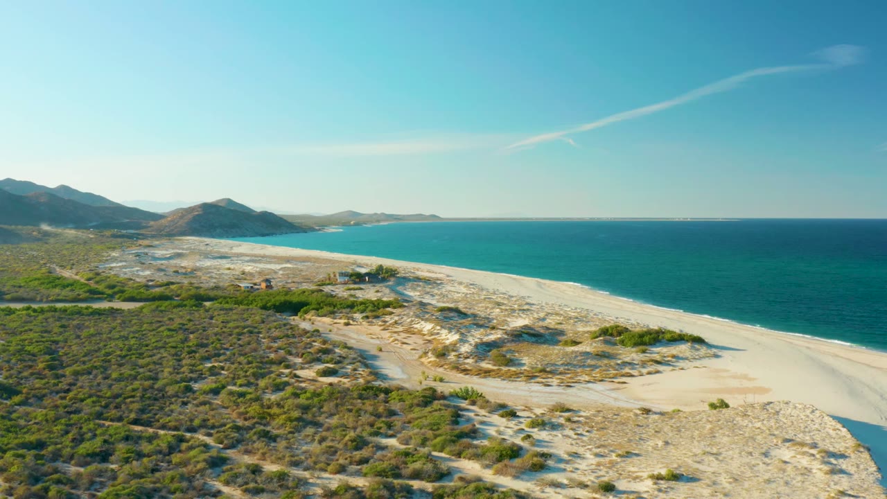 Aerial frame over Las Barracas, Baja California Sur, showing dry scrub, a sandy coastal path, pale beach, turquoise ocean, distant hills, and no visible people.