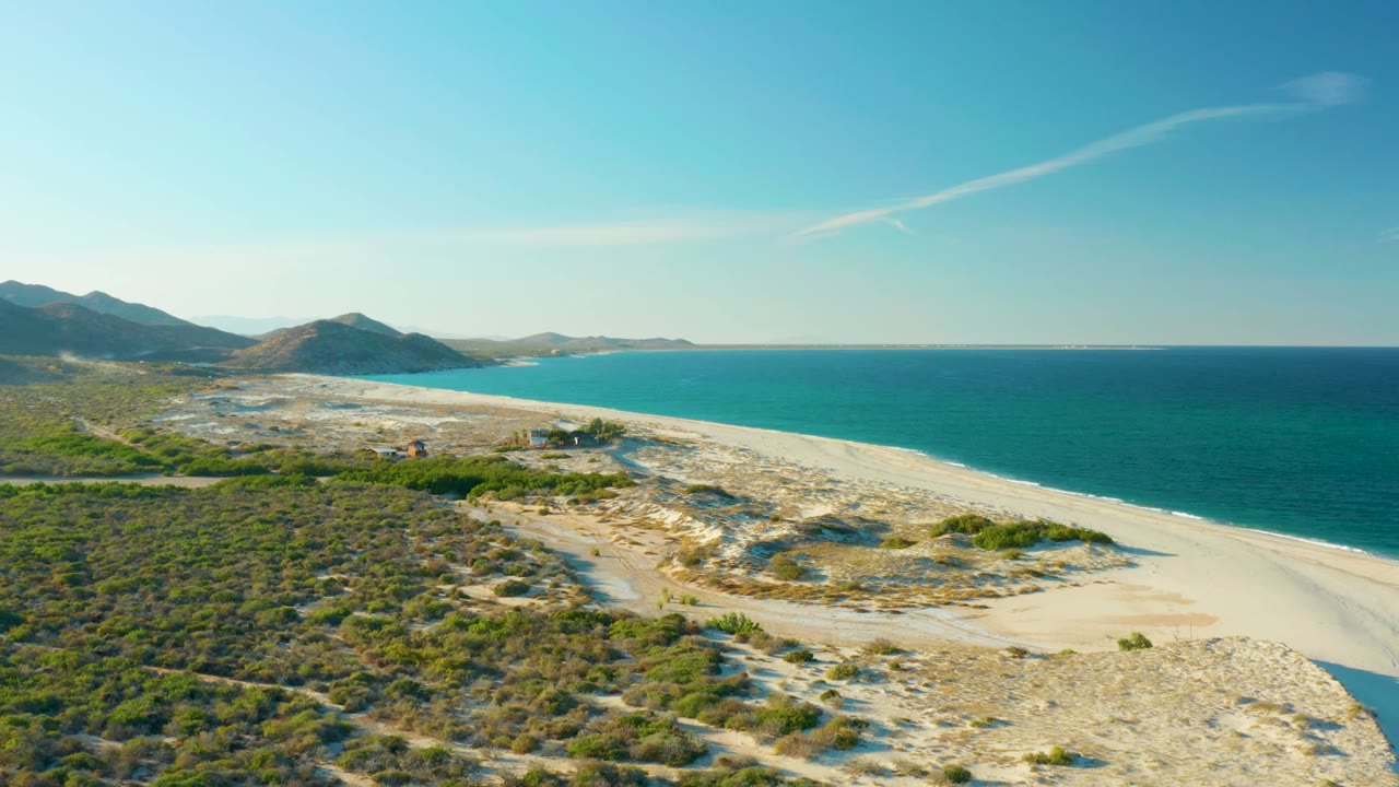 Wide aerial coast view with dirt track crossing scrub toward a sandy beach and blue ocean.