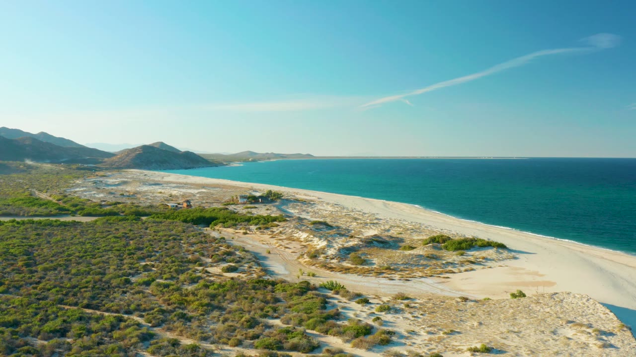 Aerial frame from Las Barracas showing scrub-covered terrain in front, a curved sandy beach, turquoise ocean to the right, low hills beyond, and no visible people.