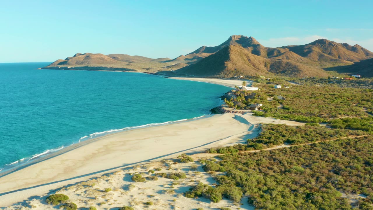 Aerial view over Las Barracas showing turquoise ocean on the left, pale sand below, green shrubs inland, distant buildings, brown mountains, and no visible people.