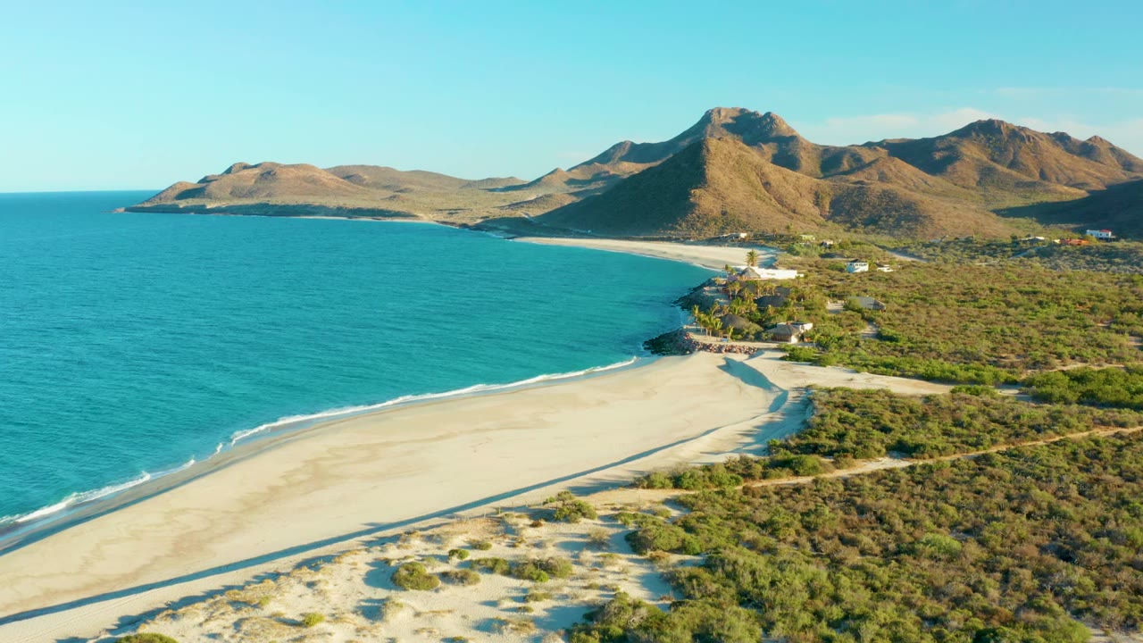 Wide aerial coastline with waves along sand and mountains inland.