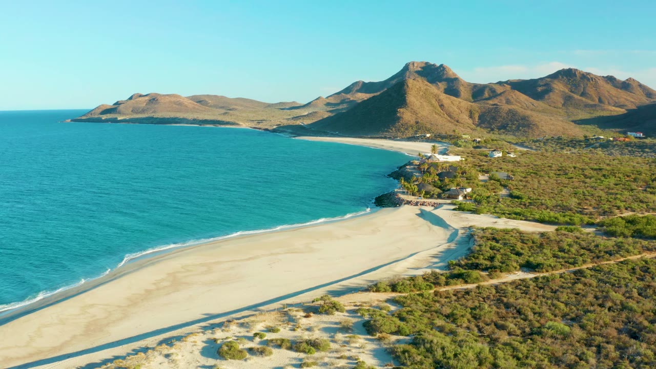 Las Barracas aerial frame with more open turquoise water, empty sand in the lower frame, dense coastal shrubs to the right, mountain slopes beyond, and no visible people.