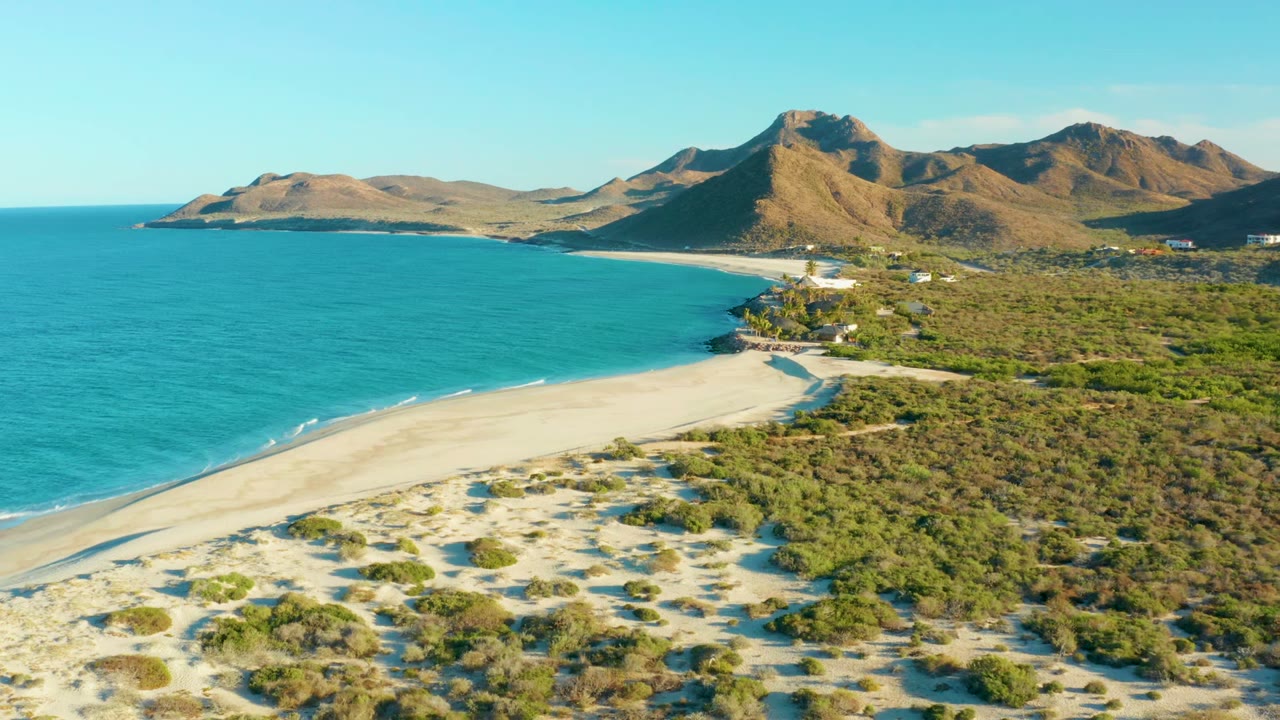 Aerial frame of Las Barracas coastline with turquoise water on the left, pale sand and scrub dunes below, brown mountains in the distance, and no visible people.