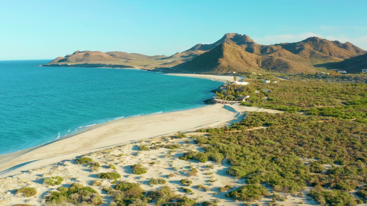 Las Barracas aerial still with turquoise ocean occupying the left side, a pale beach running diagonally through the frame, scrubland on the right, distant buildings near the shore, and no visible people.