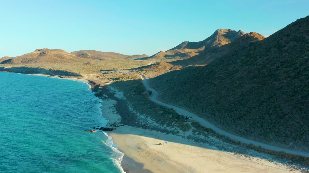 Aerial frame over Las Barracas, Baja California Sur, with turquoise water at left, a sandy beach below, a curving dirt road beside scrub hills, and distant brown mountains.