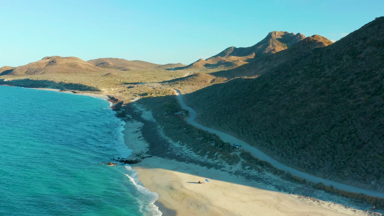 Coastal road runs between turquoise ocean and dark hillside with mountains beyond.