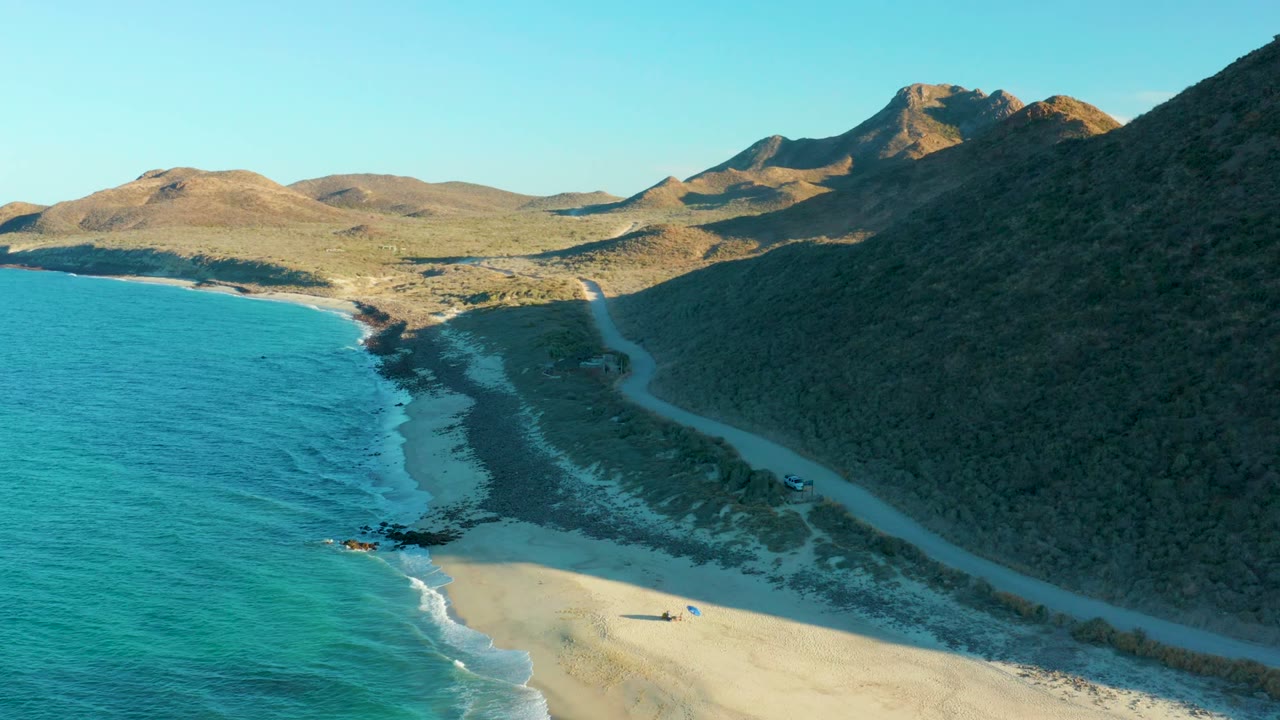 Aerial frame of the Las Barracas coast with blue water and a narrow surf line, sandy beach in the lower frame, a dirt road beside the hill, and a small vehicle near the road.