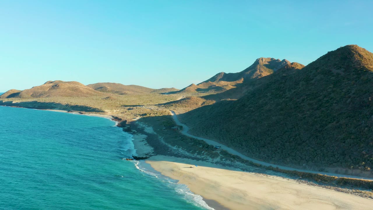 Aerial frame over Las Barracas showing turquoise ocean beside a pale sandy beach, a road along the shore, hills in the background, and no visible people.