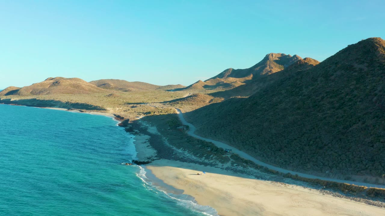 Las Barracas aerial frame with a broad blue-green ocean area, sandy beach at the lower edge, the coastal road tracking the shore, shadowed hill at right, and no visible people.