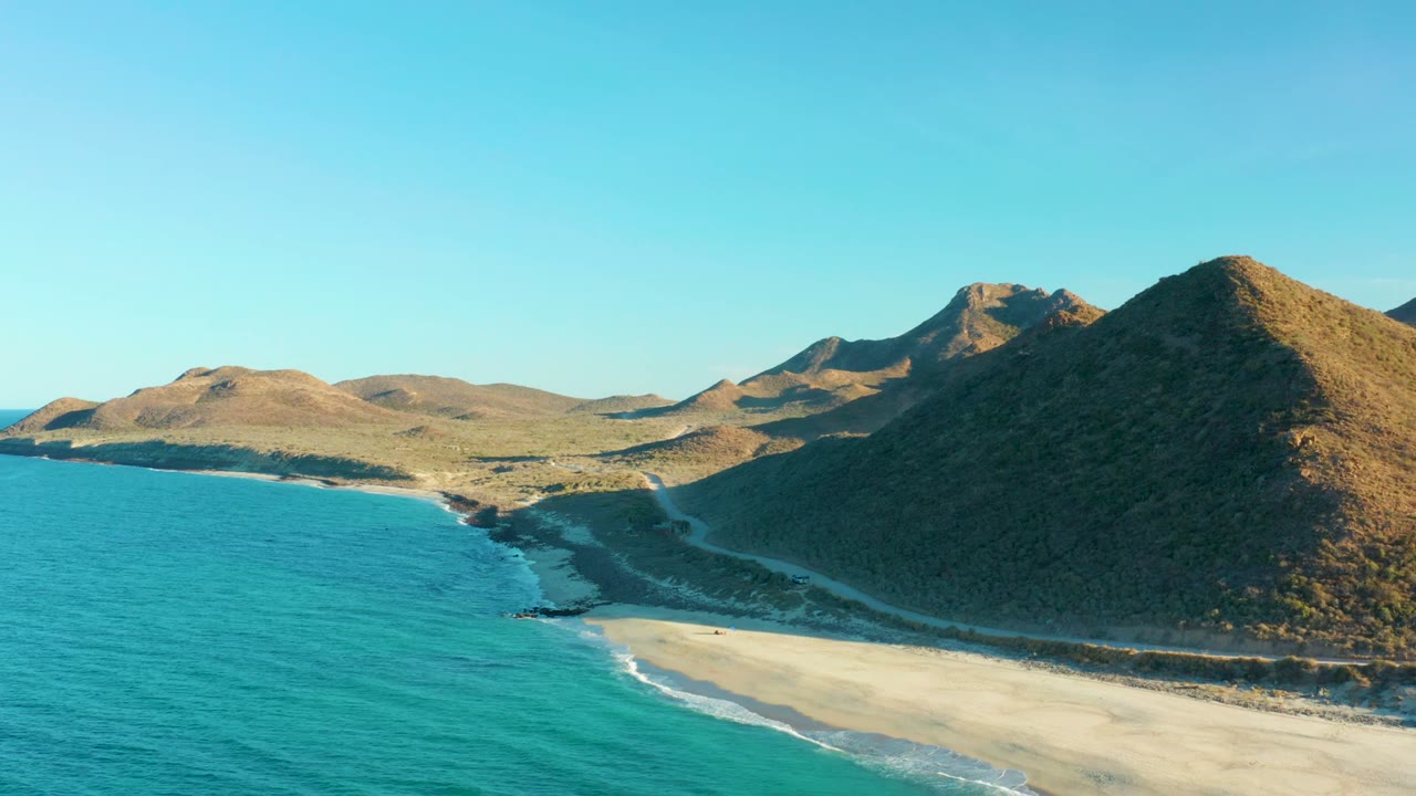 At 0:00, an aerial view over Las Barracas shows turquoise water meeting a pale sandy beach below rocky brown hills, with clear blue sky and no visible people.