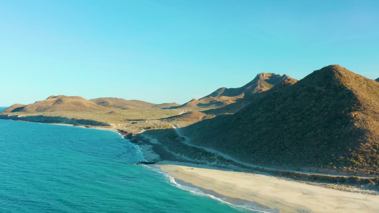 Turquoise water curves along a sandy shore below rocky hills.