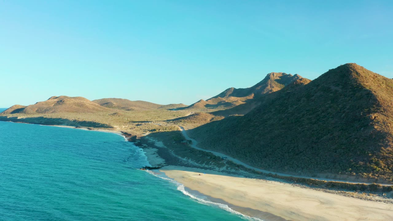 At 0:02, the Las Barracas beach runs diagonally beneath dry rocky hills, with blue-green ocean filling the left side and no visible people on the sand.