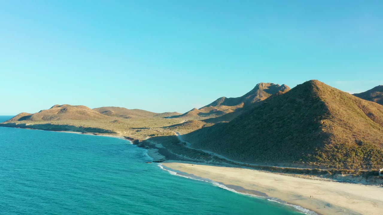 Aerial view in Las Barracas, Baja California Sur, with turquoise ocean at left, a pale sandy beach below, dry hills to the right, clear blue sky, and no visible people.