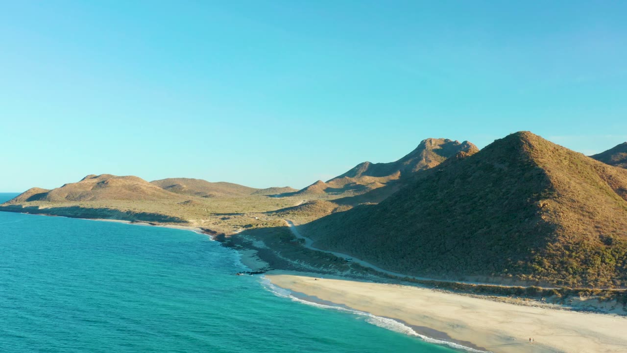 Wide aerial view over Las Barracas coastline, where teal water meets a light sand beach below rounded dry hills, with a high clear sky and no visible people.