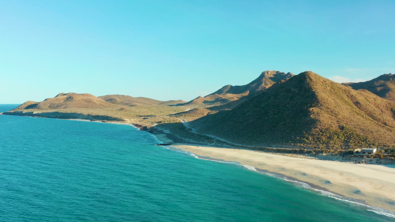 Opening frame over Las Barracas, Baja California Sur shows turquoise ocean beside a pale sandy beach with shadowed brown mountains behind it and no visible people.