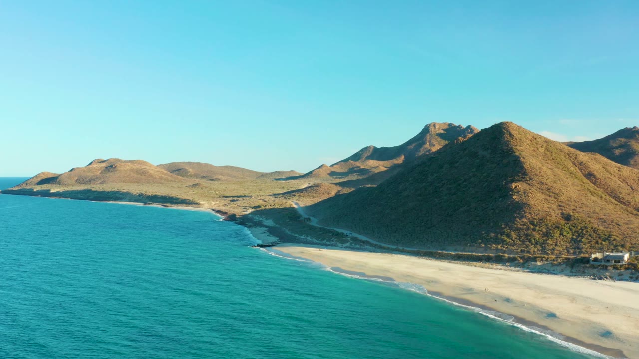 At 0:02, the Las Barracas aerial view shows more of the right shoreline edge with small structures near the beach, turquoise water in front, and no visible people.