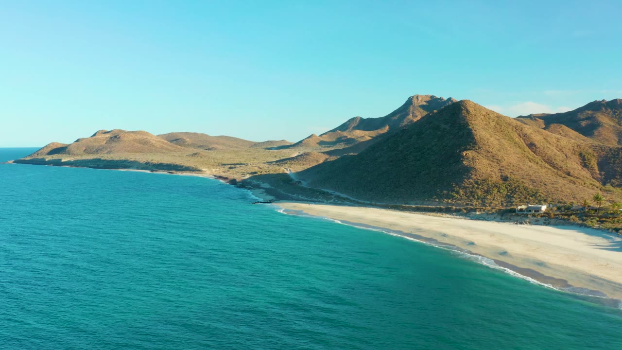 Opening aerial frame over Las Barracas with turquoise water in the foreground, a pale curving beach, dry hills beyond, and no visible people.