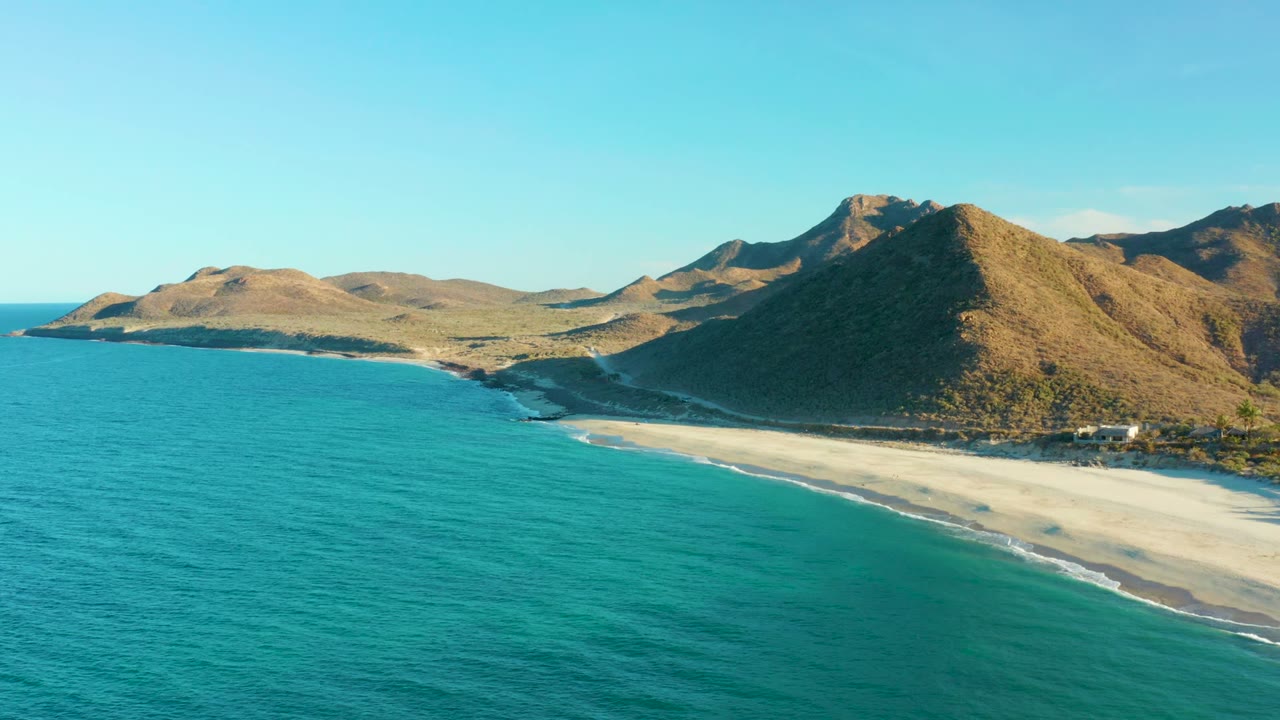 Turquoise water and a pale beach curve below sunlit hills and mountains.