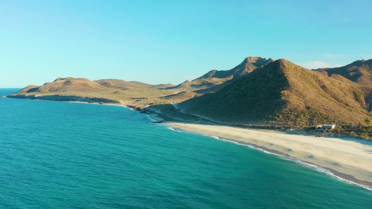 Third frame over Las Barracas with blue-green ocean occupying the foreground, a light sand beach at right, rugged hills behind it, and no visible people.
