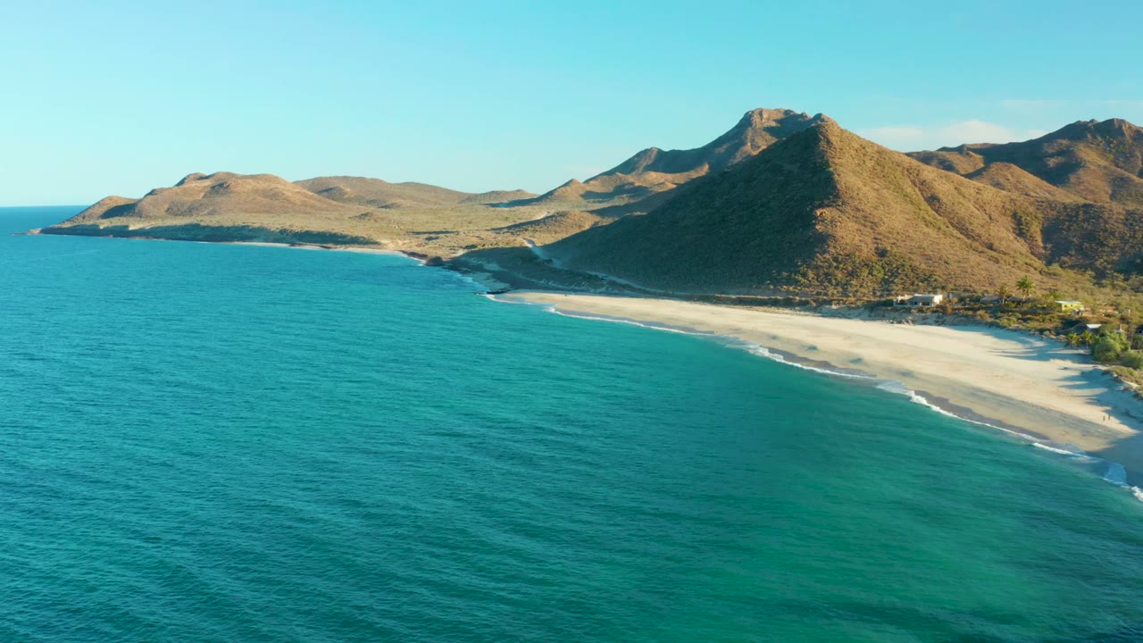 Aerial frame over Las Barracas, Baja California Sur, showing turquoise ocean before a pale sandy beach and brown mountains, with no visible people.