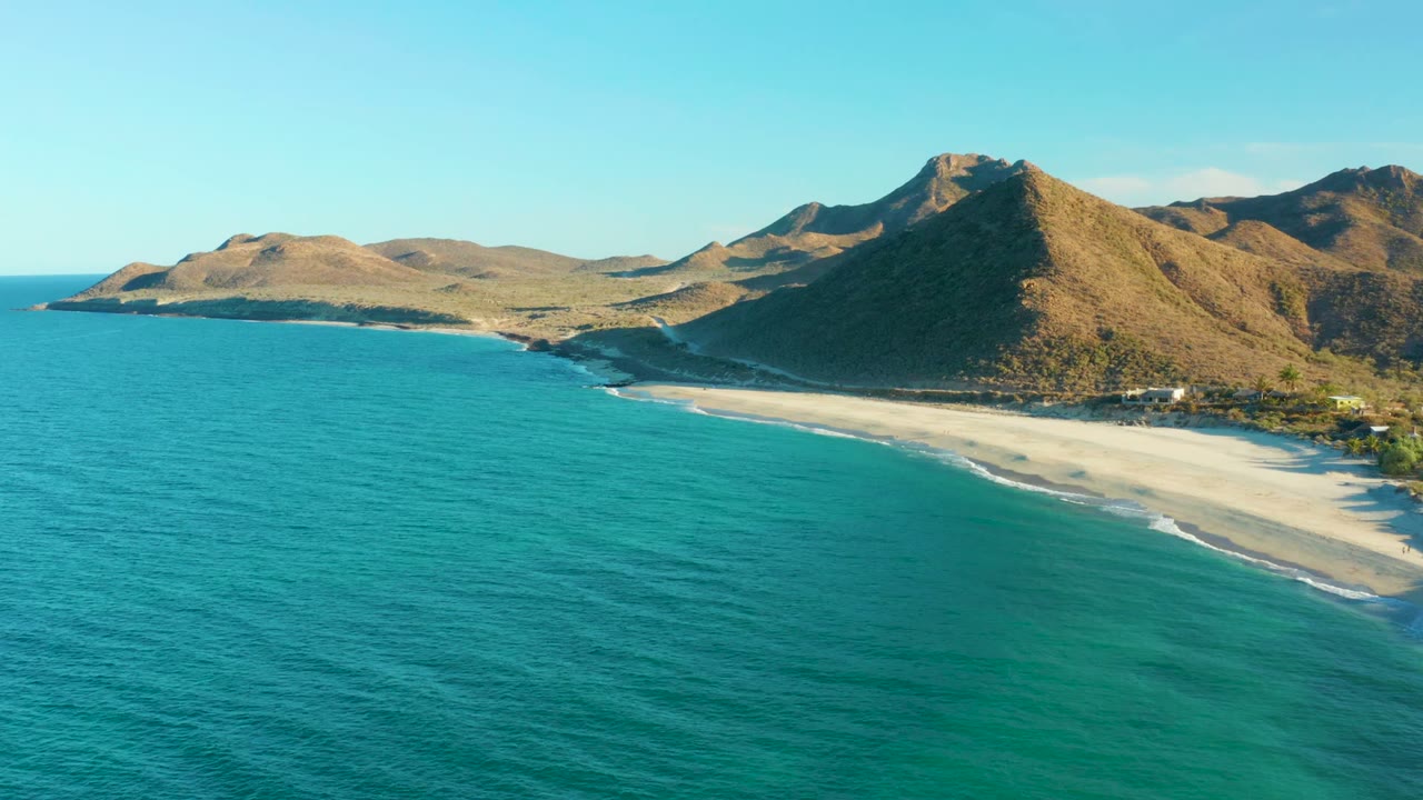 Wide aerial coastline with blue-green water, sand, and mountains under clear sky.