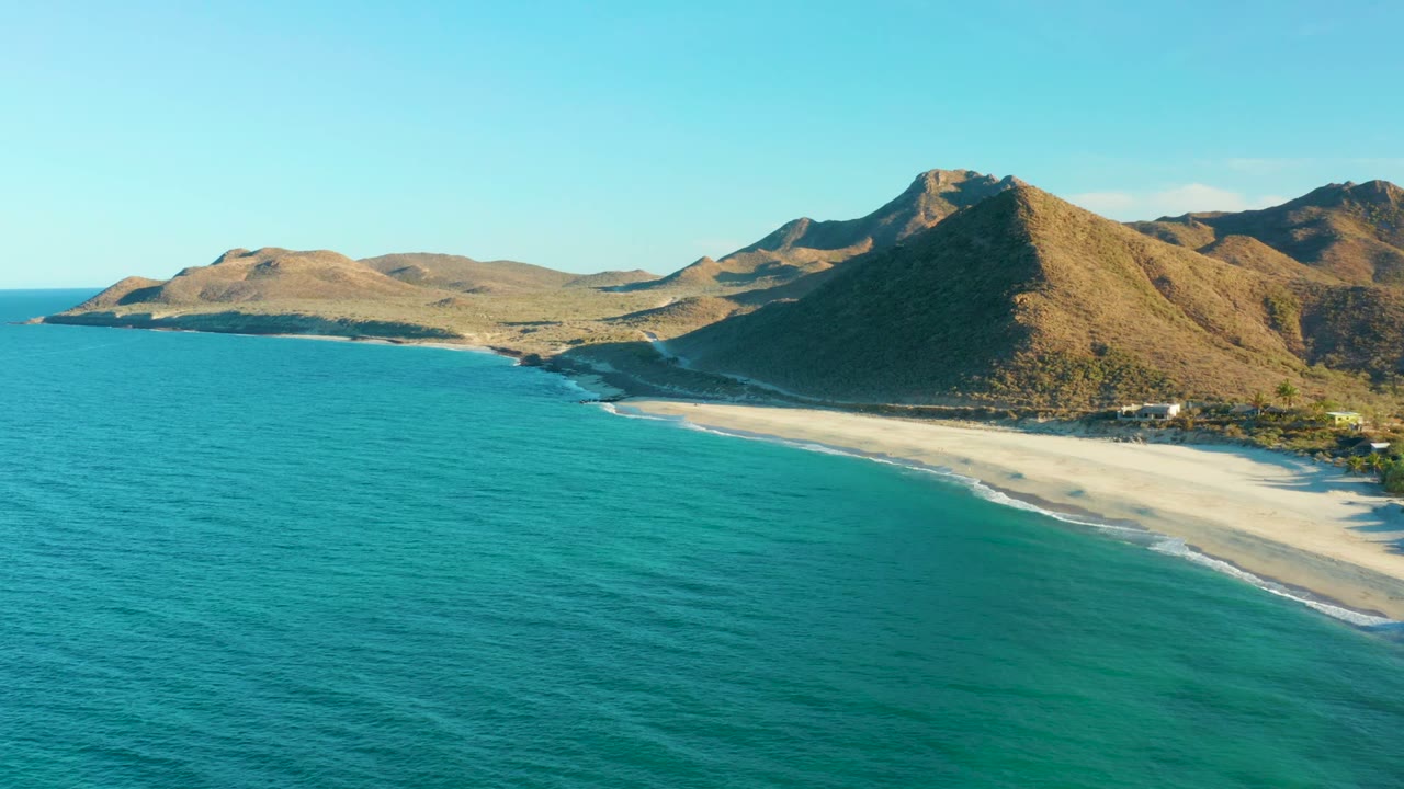 Aerial view at Las Barracas with the beach curving along turquoise water, dry mountain slopes behind it, and no visible people.