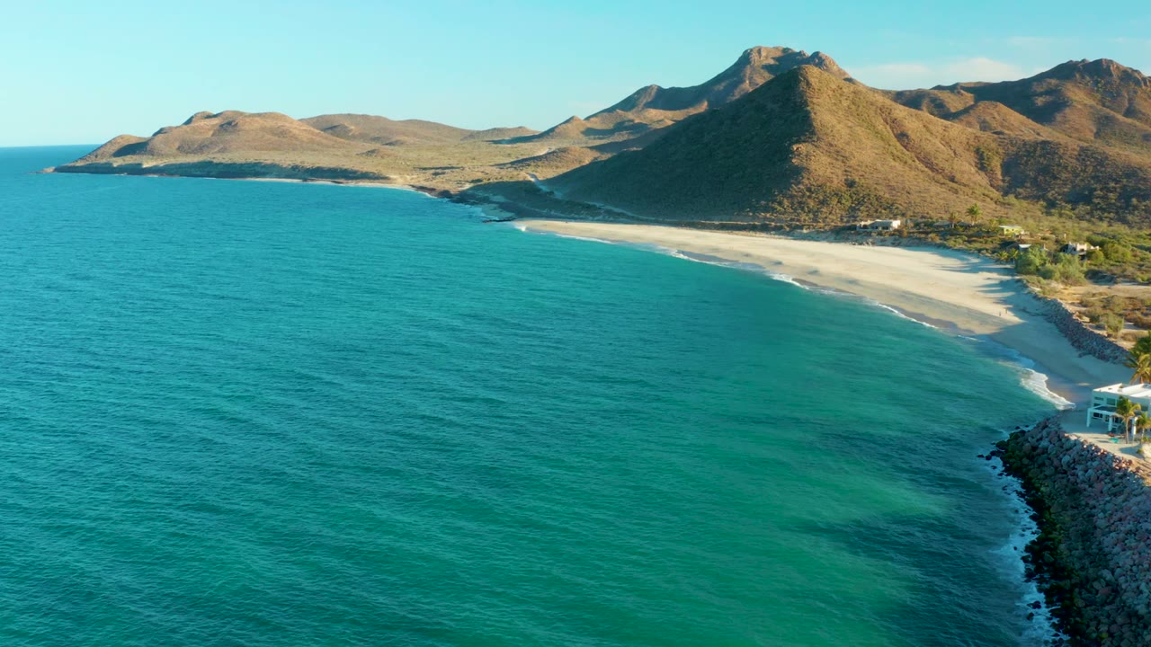 Aerial frame over Las Barracas showing turquoise water in the foreground, a pale curved beach, brown mountain ridges behind it, a rocky right edge, and no visible people.