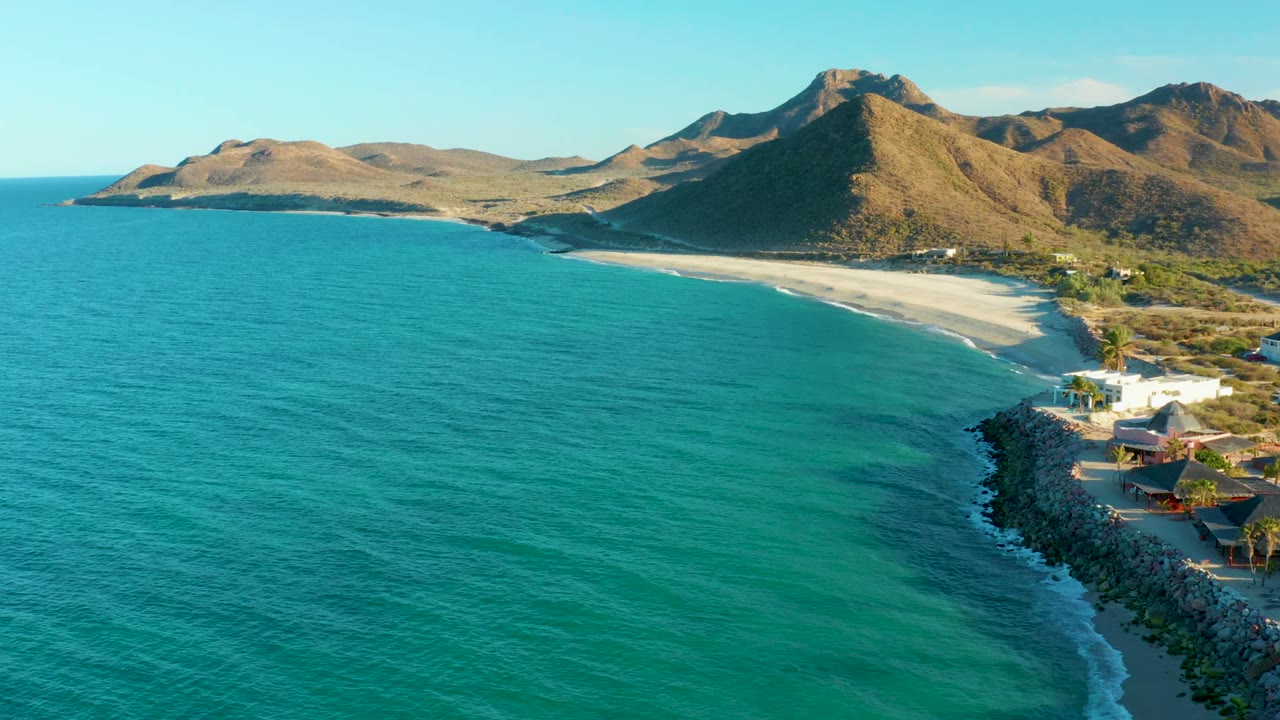 Opening aerial frame over Las Barracas, Baja California Sur, with turquoise ocean in front, a curved sandy beach below brown mountains, small buildings at right, and no visible people.