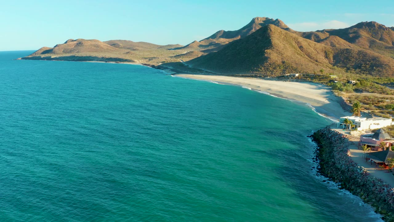 Mid-clip aerial frame at Las Barracas showing turquoise sea across most of the image, a sandy beach backed by dry hills, a rocky point with buildings at right, and no visible people.