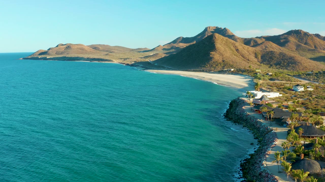 Opening aerial frame over Las Barracas shows turquoise ocean, a rocky shore at right, a pale beach below dry mountains, scattered coastal buildings, palm trees, and no visible people.