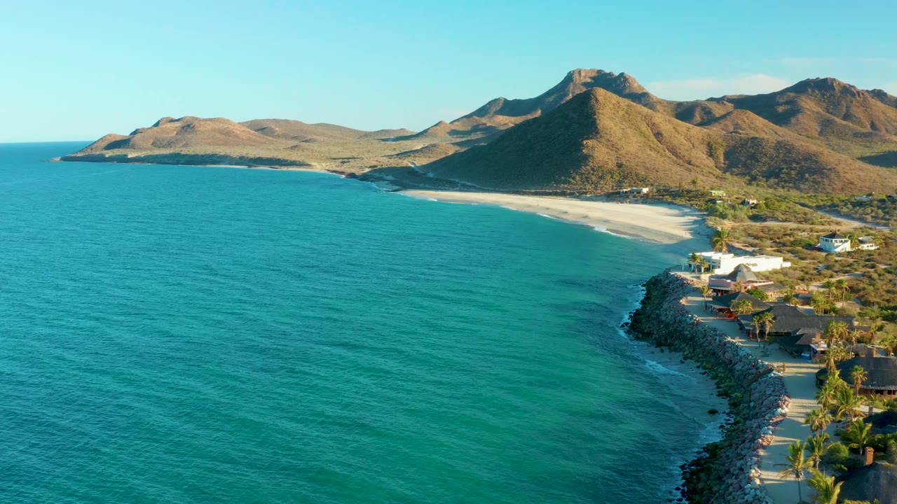 Turquoise water and rocky shore lead toward a sandy beach and dry mountains under clear sky.