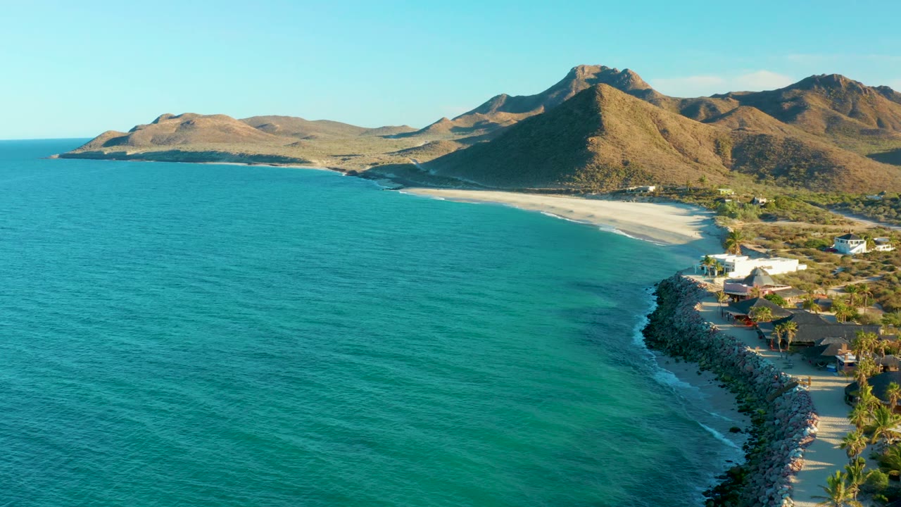At 0:02, the Las Barracas shoreline remains at frame right with blue-green ocean in front, a curved sandy beach beyond it, dry mountains behind, coastal buildings, palms, and no visible people.