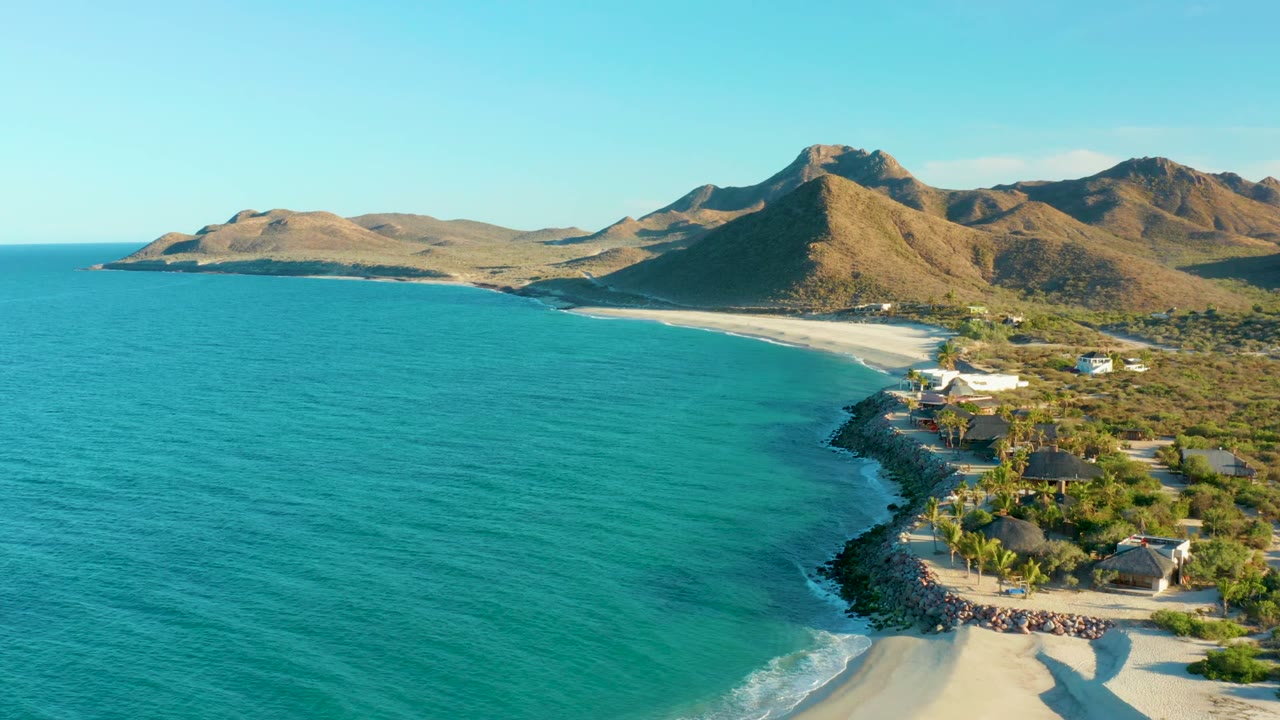 Aerial frame over Las Barracas showing turquoise ocean in the foreground, a curved sandy beach, shoreline buildings, dry mountains, and tiny people near the waterline.