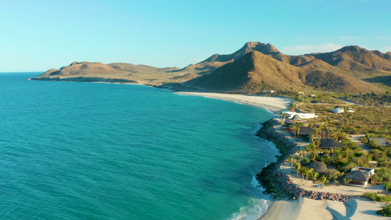 Aerial view at 0:01 over Las Barracas with blue-green ocean covering most of the frame, pale sand curving below dry hills, and beach structures clustered at the right edge.