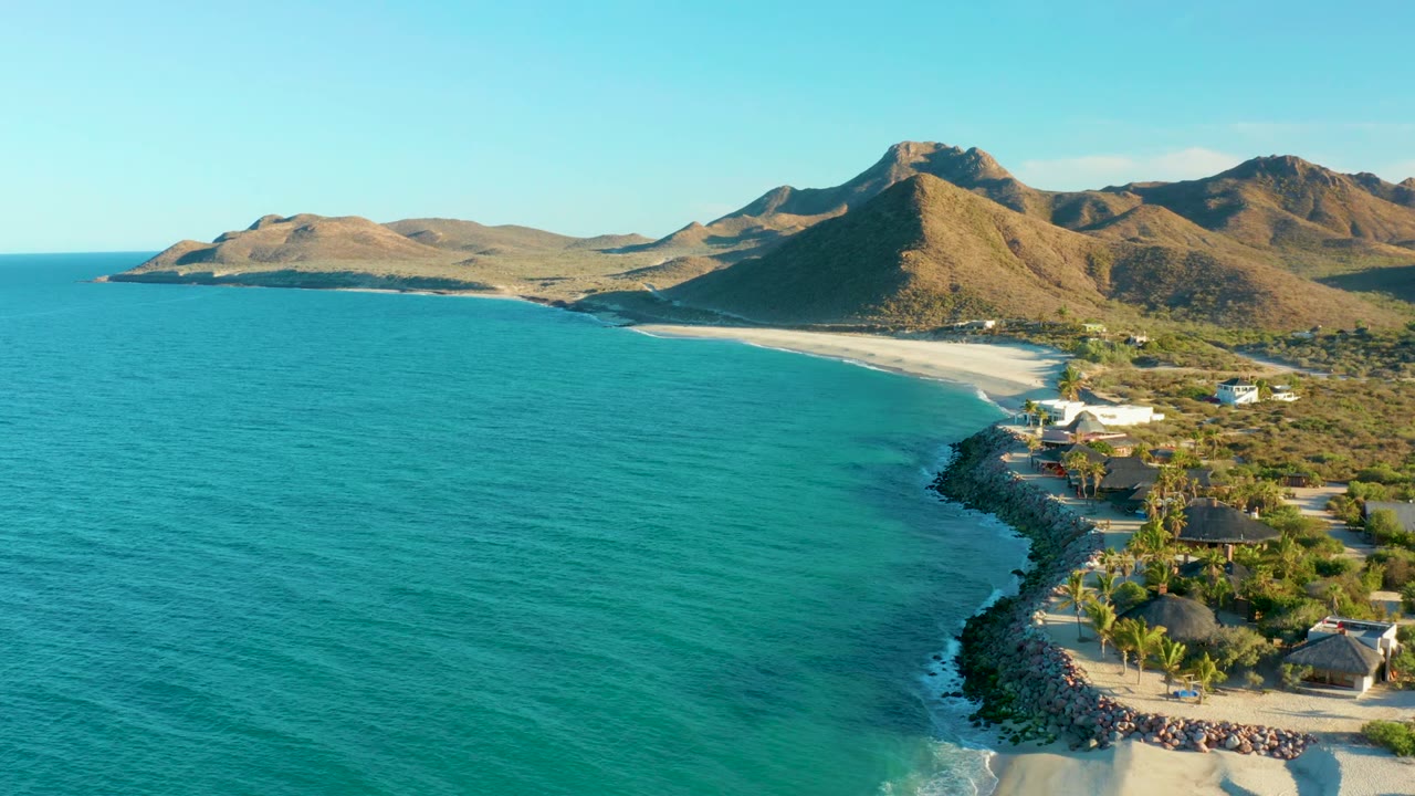 Aerial frame over Las Barracas where the rocky shoreline and thatched roofs sit at lower right, turquoise water fills the center, and brown mountains run along the horizon.