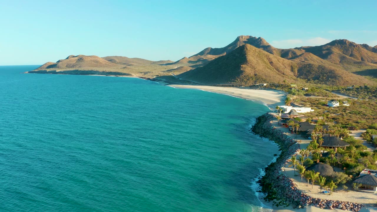 Drone view of calm ocean, sandy shoreline, small buildings, and mountains.