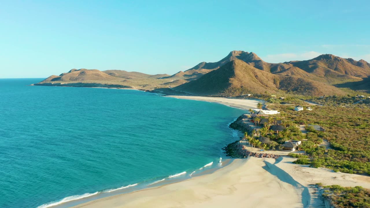 Aerial view over Las Barracas with turquoise ocean on the left, pale sand in the foreground, coastal buildings and scrubland on the right, mountains in the background, and no visible people.