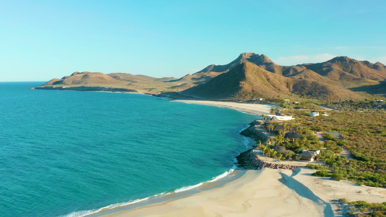 Drone view of beach, surf line, coastal vegetation, buildings, and mountains.