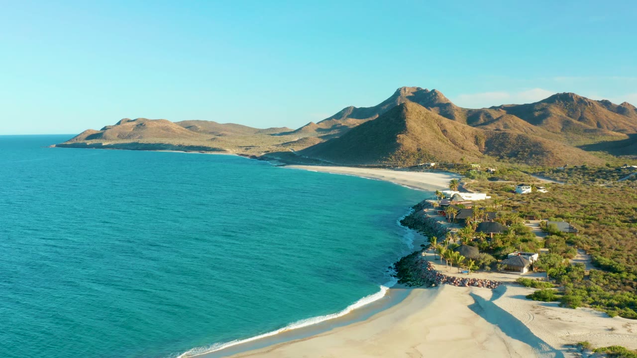 Aerial view at Las Barracas with more open turquoise water filling the left side, a sandy beach and rocky shoreline below, coastal structures to the right, mountains beyond, and no visible people.