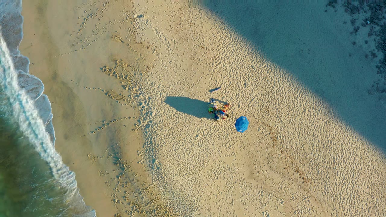 Overhead frame from Las Barracas, Baja California Sur, showing a person lying on pale sand near a blue umbrella while white surf borders the left side and a broad shadow crosses the upper beach.