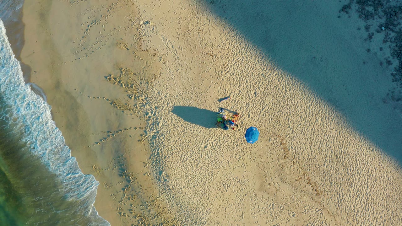 Aerial view of a beachgoer and blue umbrella as waves recede along the shoreline.