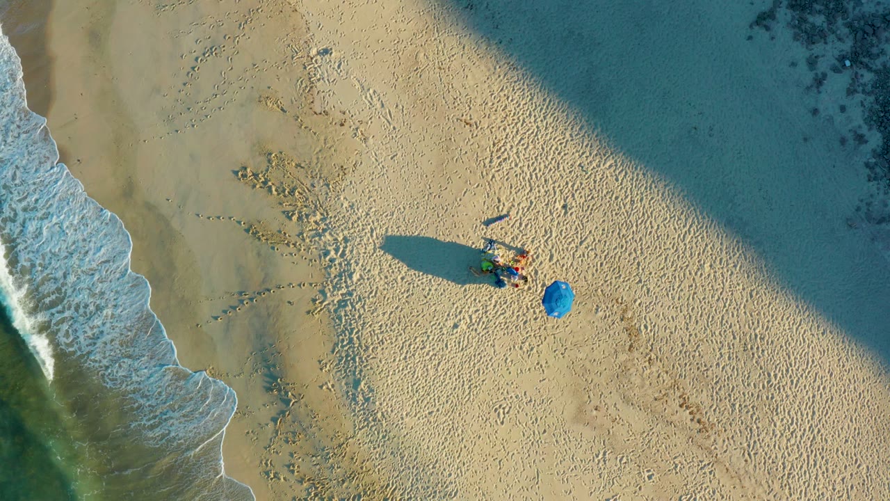 At 0:02, the Las Barracas shoreline appears from above with turquoise water and foam at the right edge, a beachgoer on the sand, a separate blue umbrella, and scattered footprints nearby.