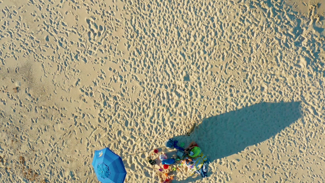 Top-down view of Las Barracas sand with a blue umbrella left of colorful beach gear, many footprints, a long shadow extending across the frame, and no visible people.