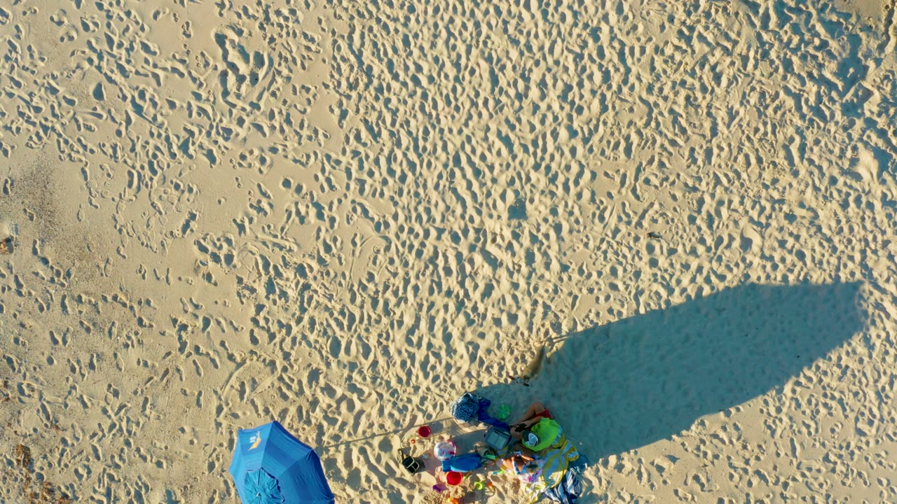 Overhead sand scene with umbrella, beach items, footprints, and large shadow slightly shifted.