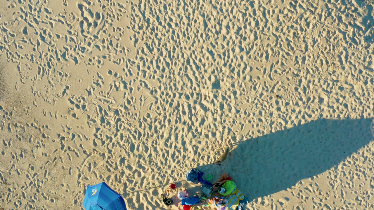 Top-down Las Barracas beach frame showing rippled sand, dense footprints, a blue umbrella at lower left, colorful gear to the right, and a broad shadow falling across the sand with no visible people.