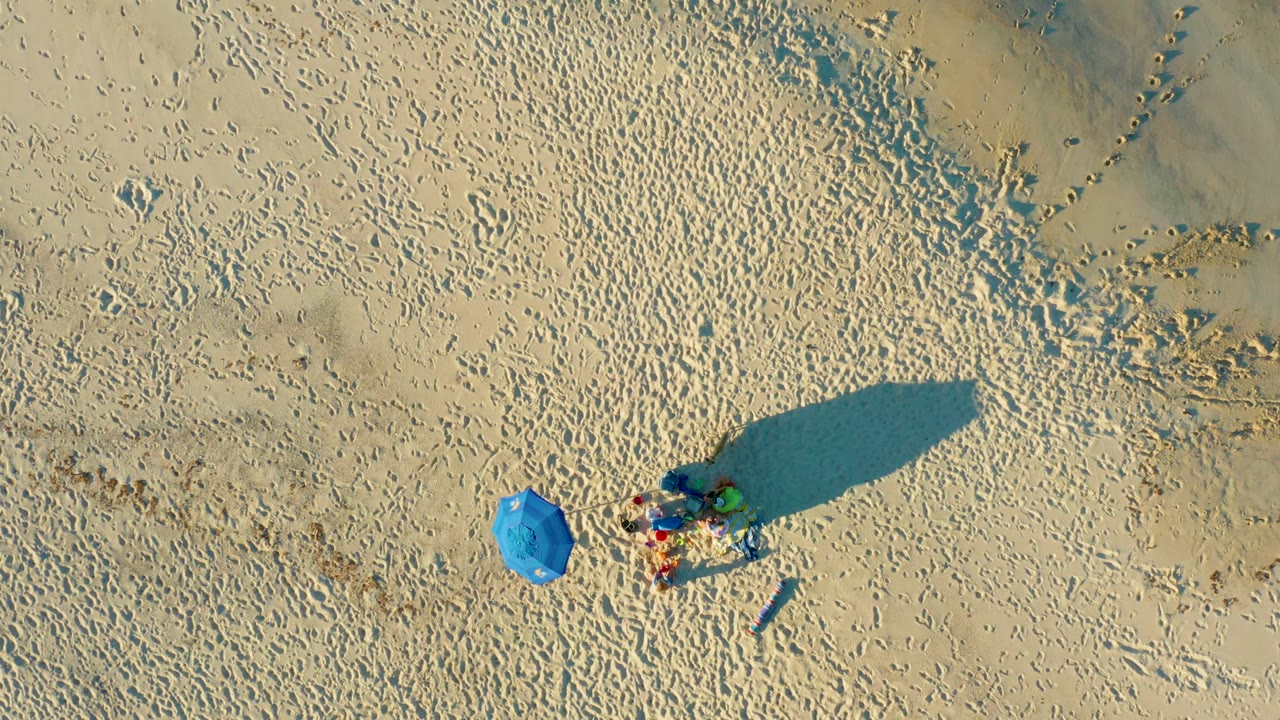 Overhead view in Las Barracas, Baja California Sur, showing a blue umbrella left of a small group of people on footprint-marked sand with long shadows.